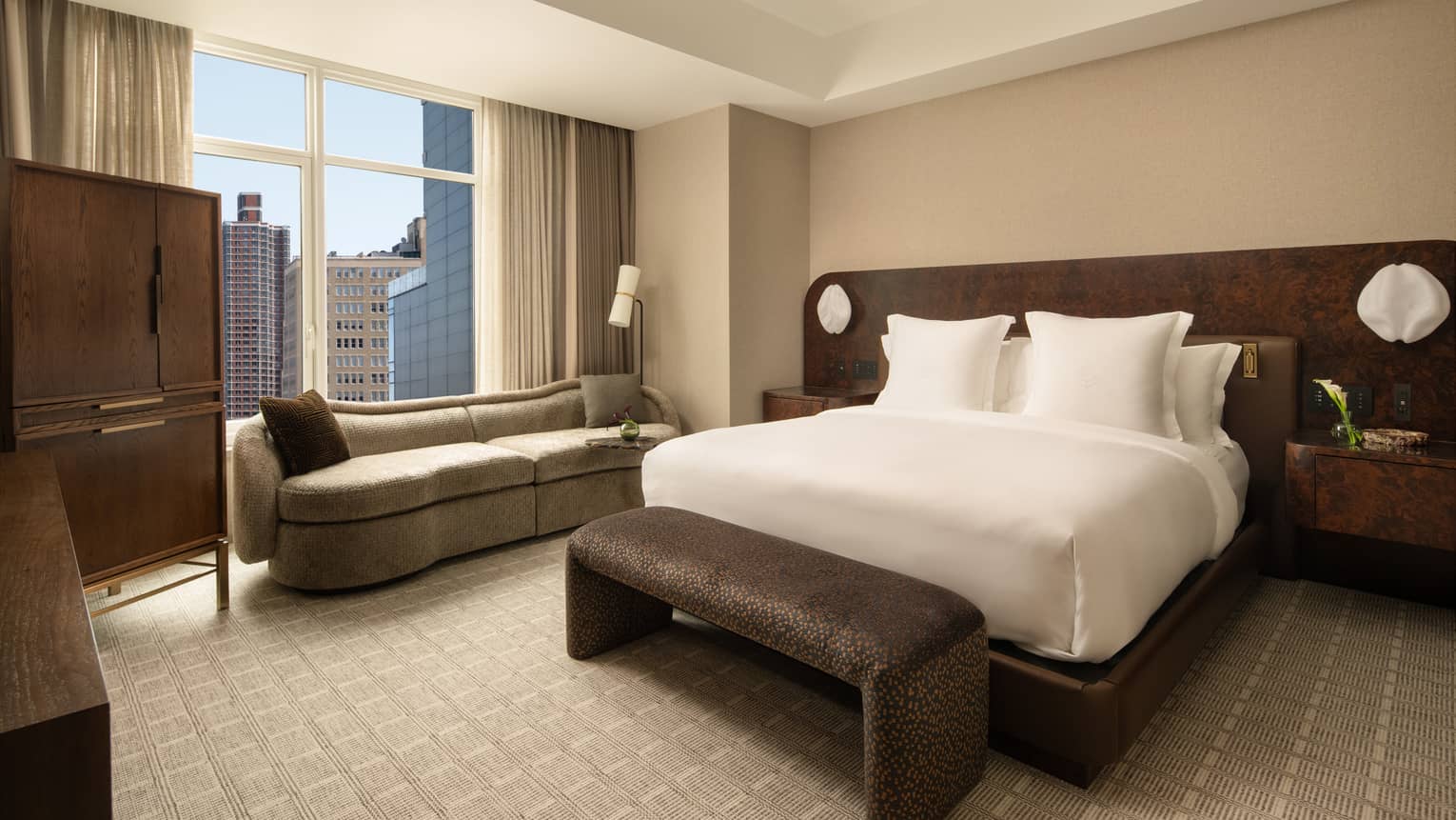 Contemporary bedroom of hotel suite, featuring a king bed, windowside chaise, and cushioned bench