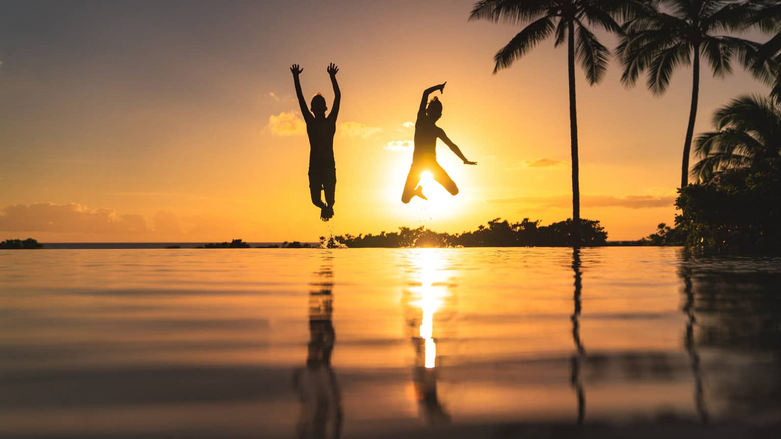 Silhouette of two children jumping above the water, sunset and palm trees in background 