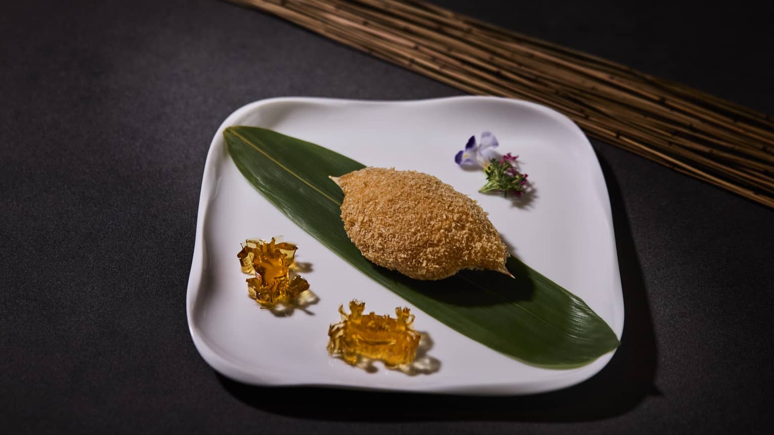 A square white plate holds a textured light-brown food item placed on an elongated dark-green leaf, surrounded by garnishes.
