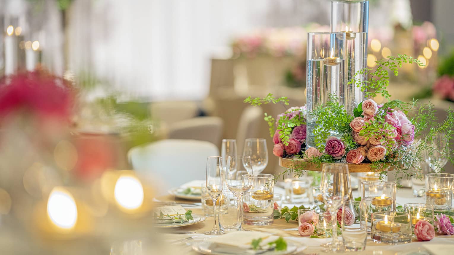 A wedding table setting featuring soft flickering candles in long glass holders, pink roses centerpiece and champagne glasses