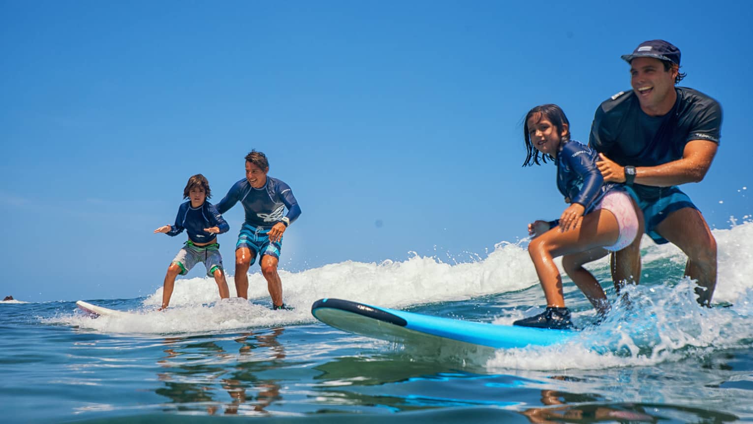 A pair of smiling adults and children standing on two surfboards, wearing swimsuits and rash guards, riding a small wave.