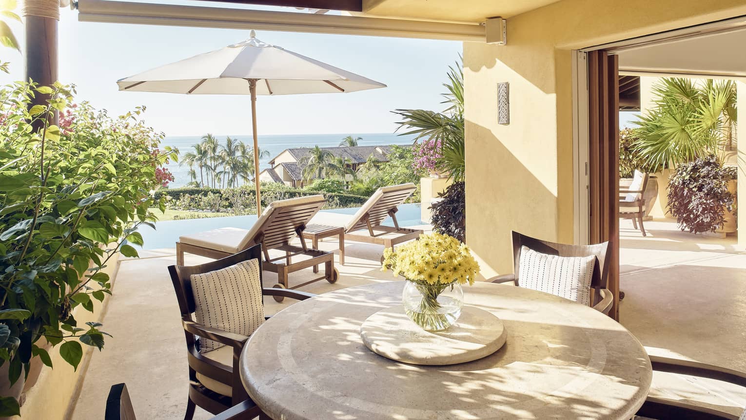 Round dining table with yellow flowers in vase under covered patio, umbrella in sun