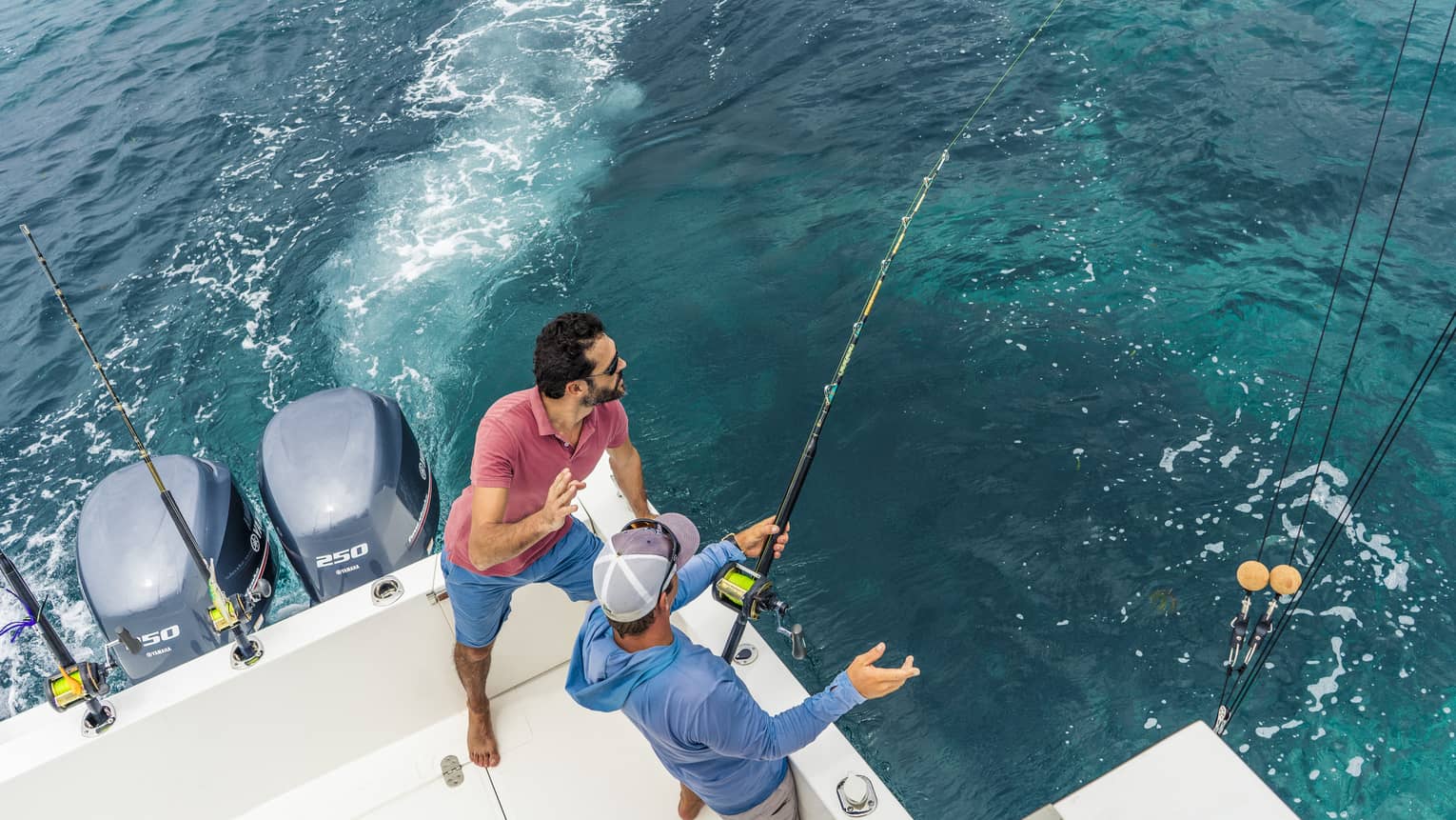Aerial view of two people fishing off the back corner of a motorboat. One grips a fishing rod while the other looks on.