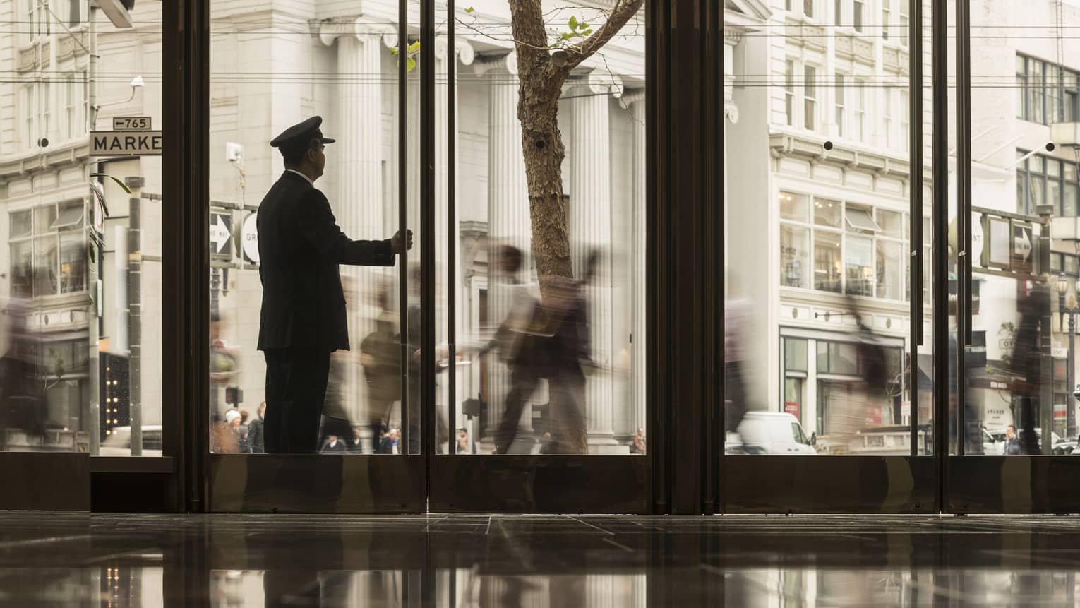 A doorman holding the door of a hotel lobby while looking outside at people walking around the city.