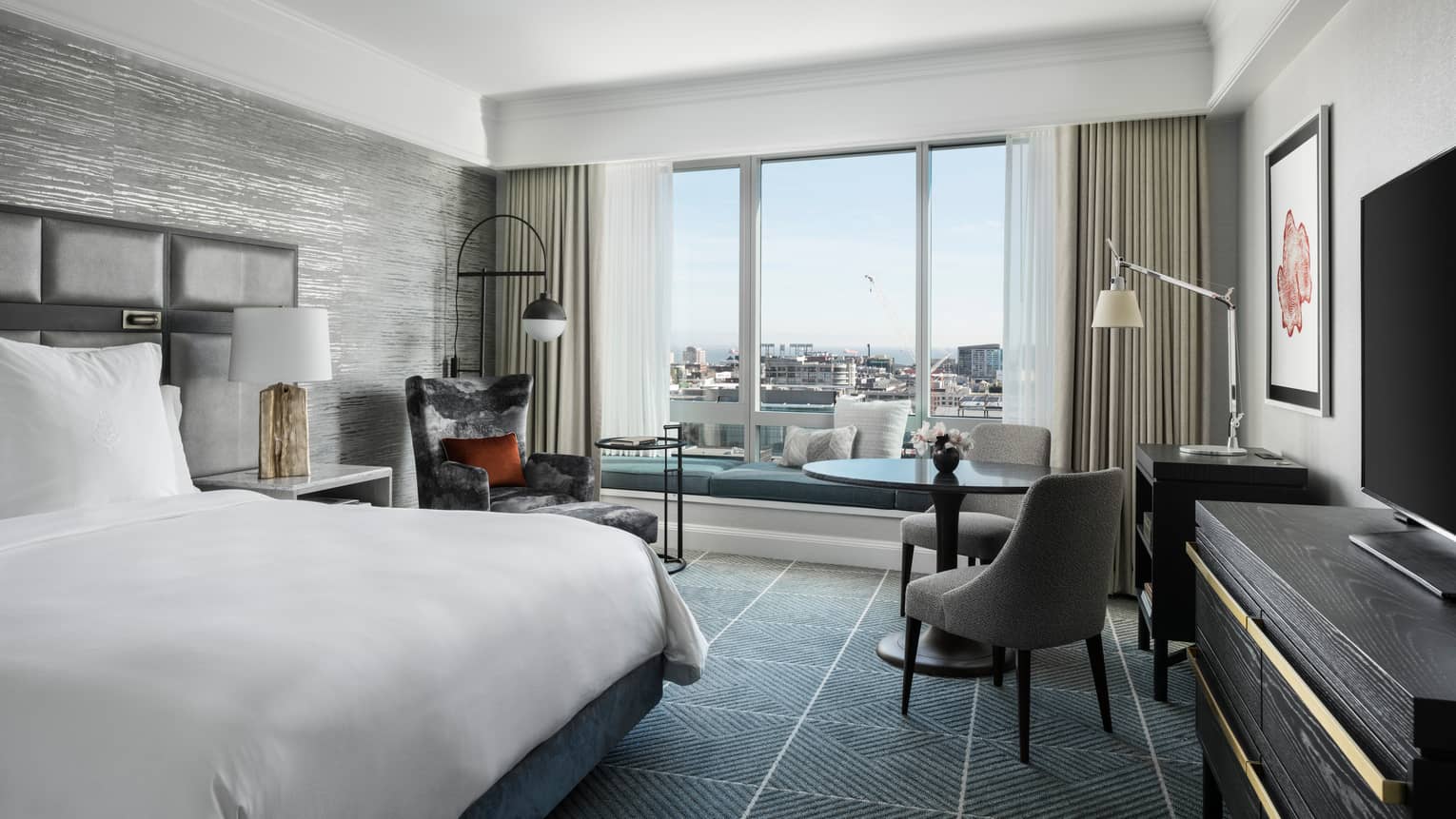 Modern hotel room with king bed, table and two upholstered grey chairs, windows overlooking San Francisco