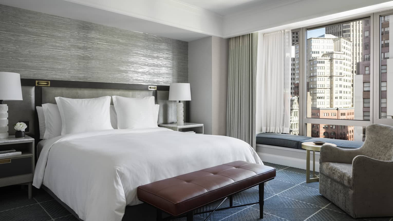 A hotel room with a bed with white sheets, a red bench, a grey chair, two white lamps, and a large window looking out at a city.