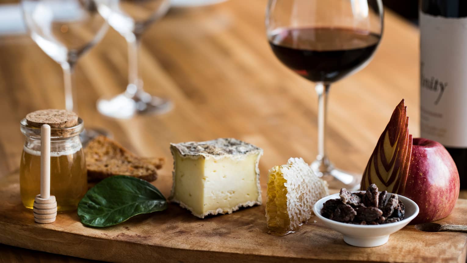 A glass of red wine with a luxurious wooden tray holding cheese, honeycomb, honey, sliced apple, nuts and crackers.