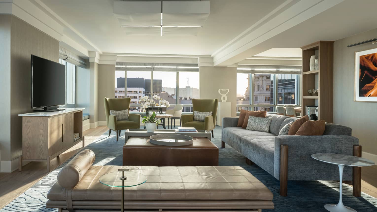 Living room with table, grey sofa, two green arm chairs, TV, chaise, windows overlooking city