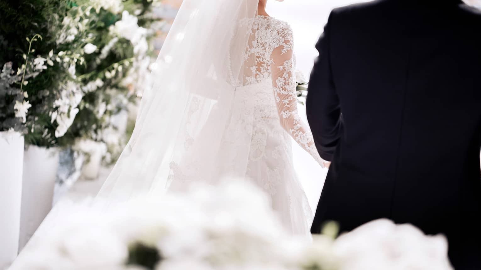 A bride in a white, lace, long sleeve gown smiles as she walks down the aisle with her groom. They are surrounded by artfully arranged white flowers.