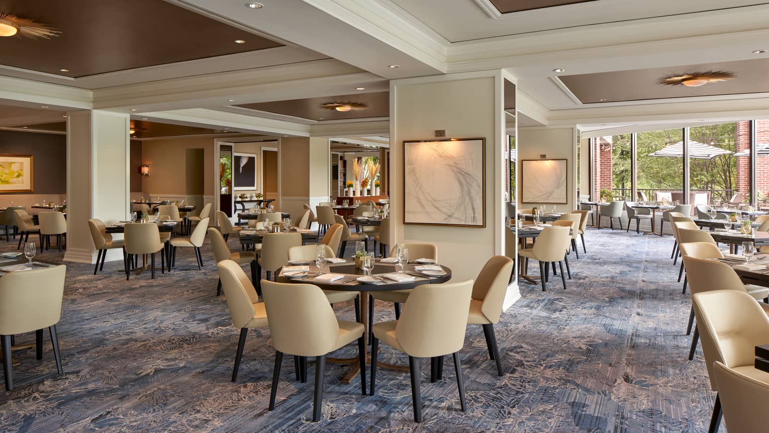 A well lit restaurant with beige chairs and round tables and large open windows.