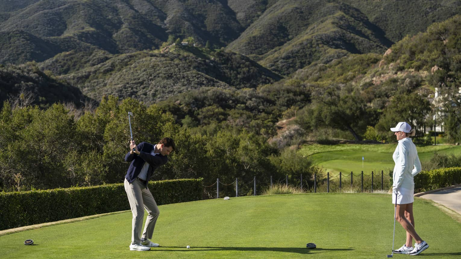 A man and woman playing golf on the green with green covered mountains in the distance.