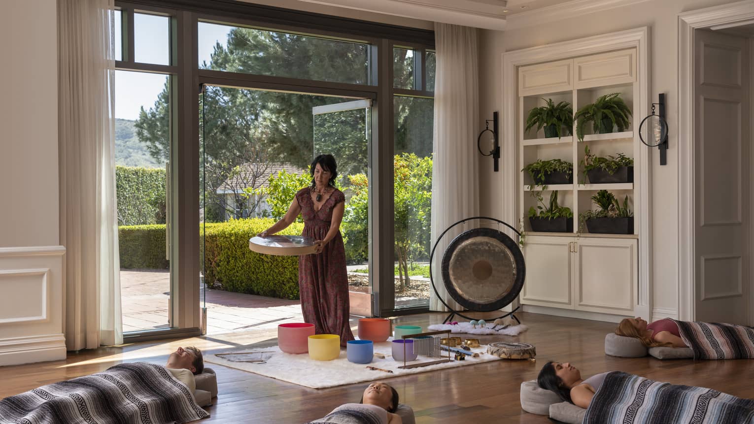 A woman standing over colourful buckets with people laying on the floor around her, there is a gong and other instruments.