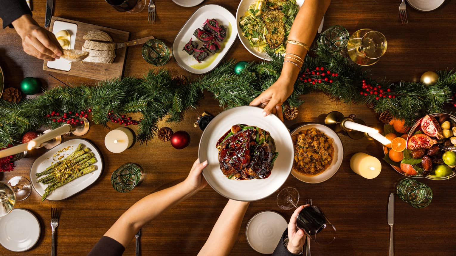 Hands pass a dish of food to another over a table set for a holiday meal with a green garland in the centre