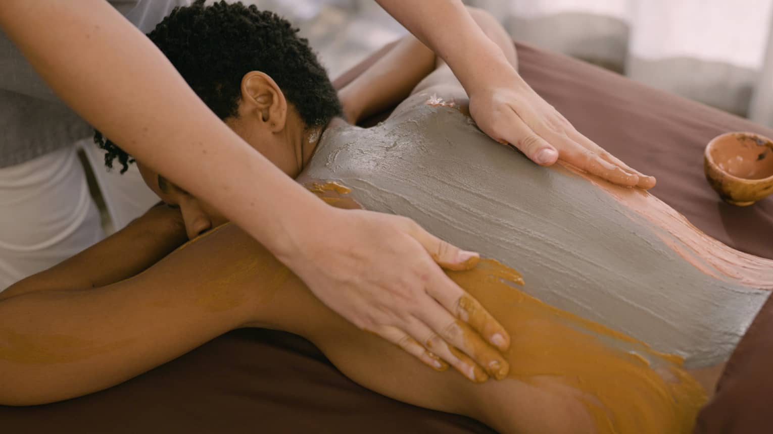 Hands rub clay across person's back as they lay on massage table