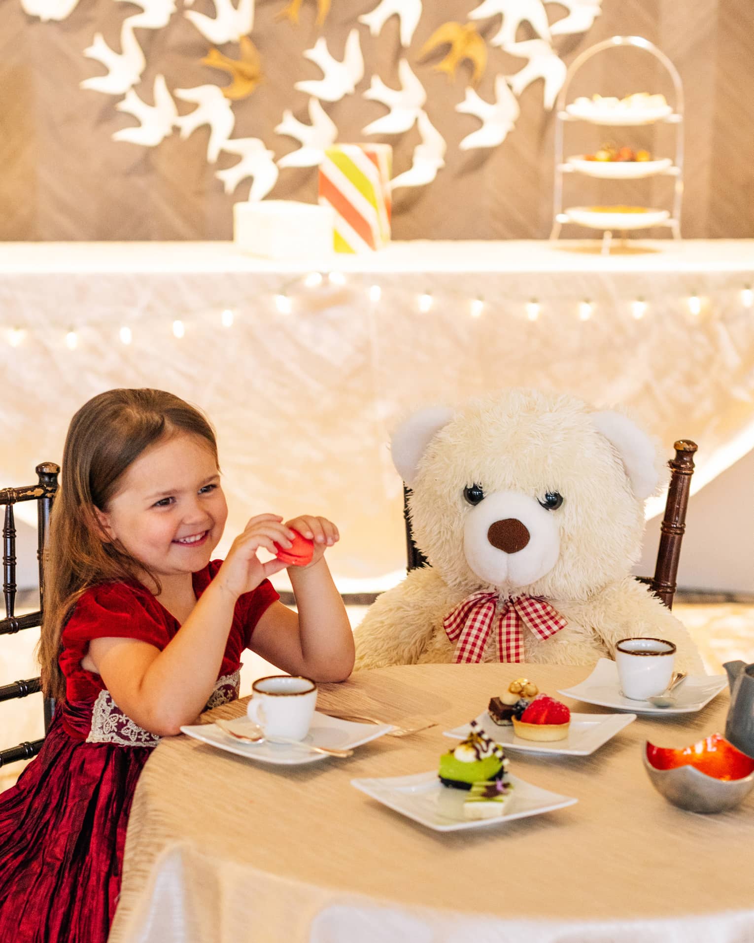 A small girl in a red dress sitting with a large teddy bear at a table with tea and small desserts.