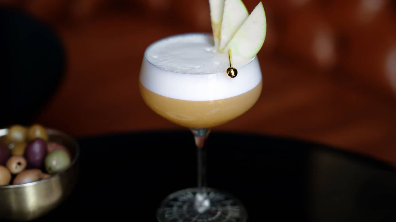 A clear stemmed glass holds a light brown drink with a white layer on top, garnished with sliced fruit.