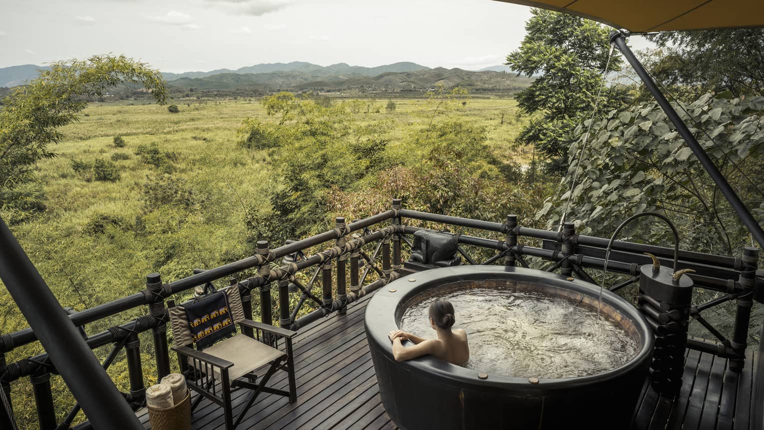 Woman wades in large round hot tub on wood deck overlooking green field, mountains