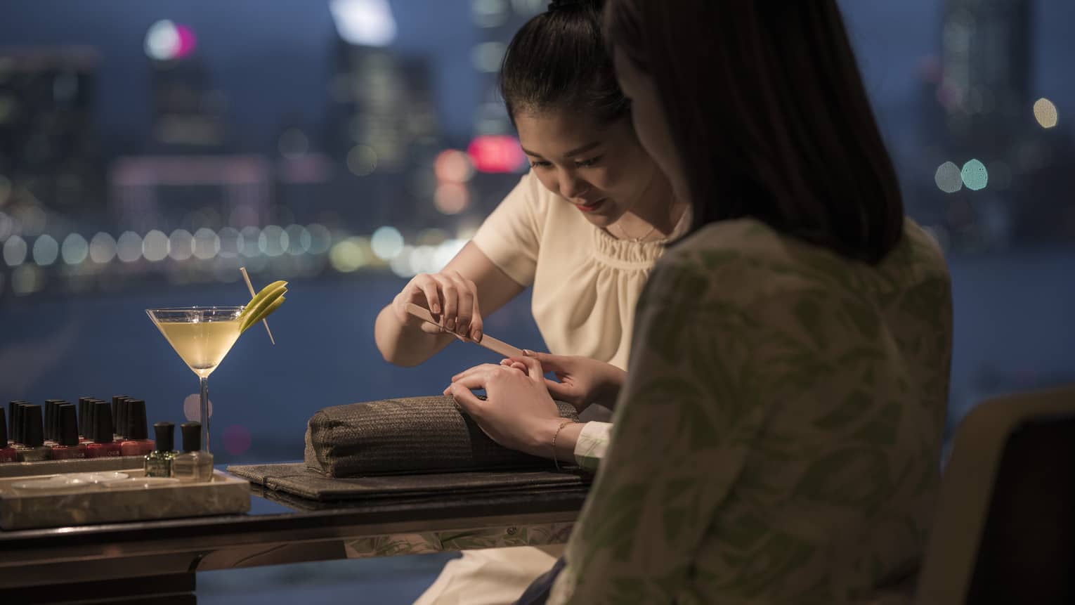 A person gets a manicure at a spa at night, overlooking a harbor. A martini sits on the table beside their hand, with nail polishes in the background.
