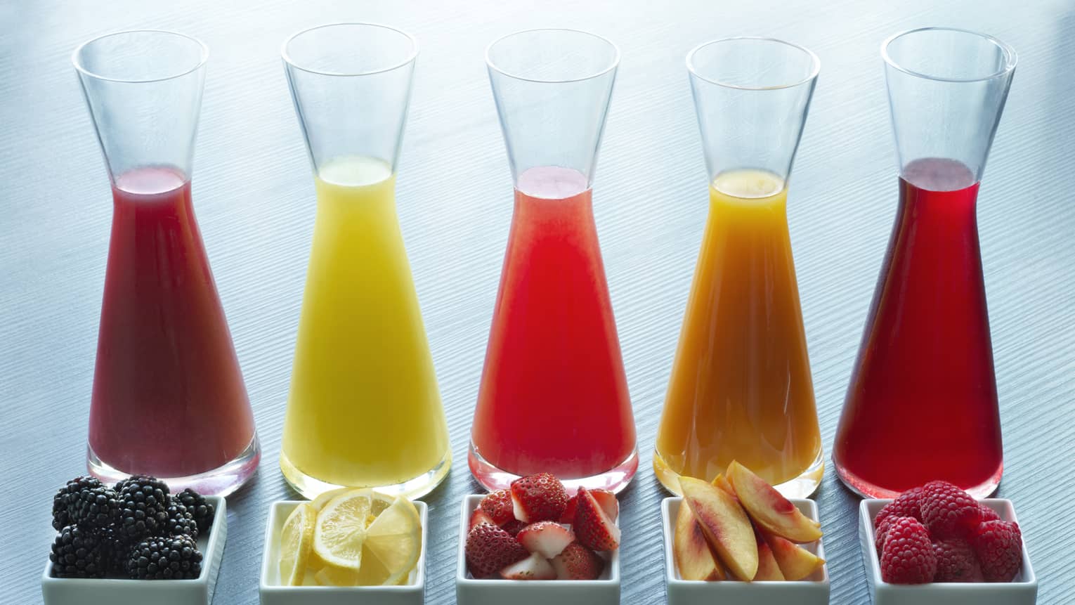Five glass juice jugs aligned with five small trays of fruit