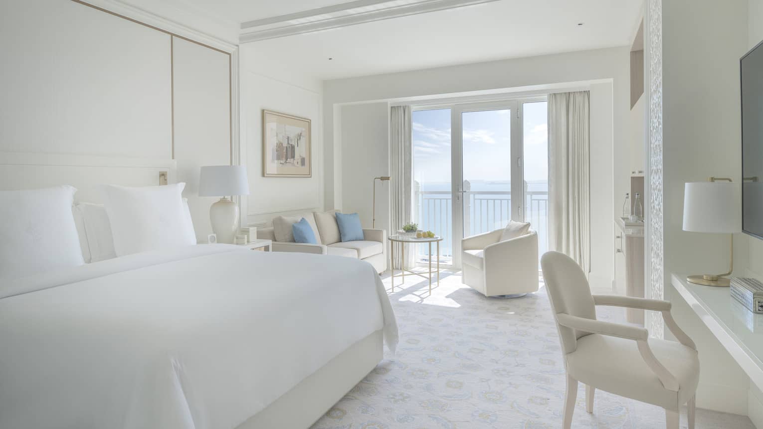 Bedroom with white bed, white walls and a white sofa and arm chair, with balcony offering sea view
