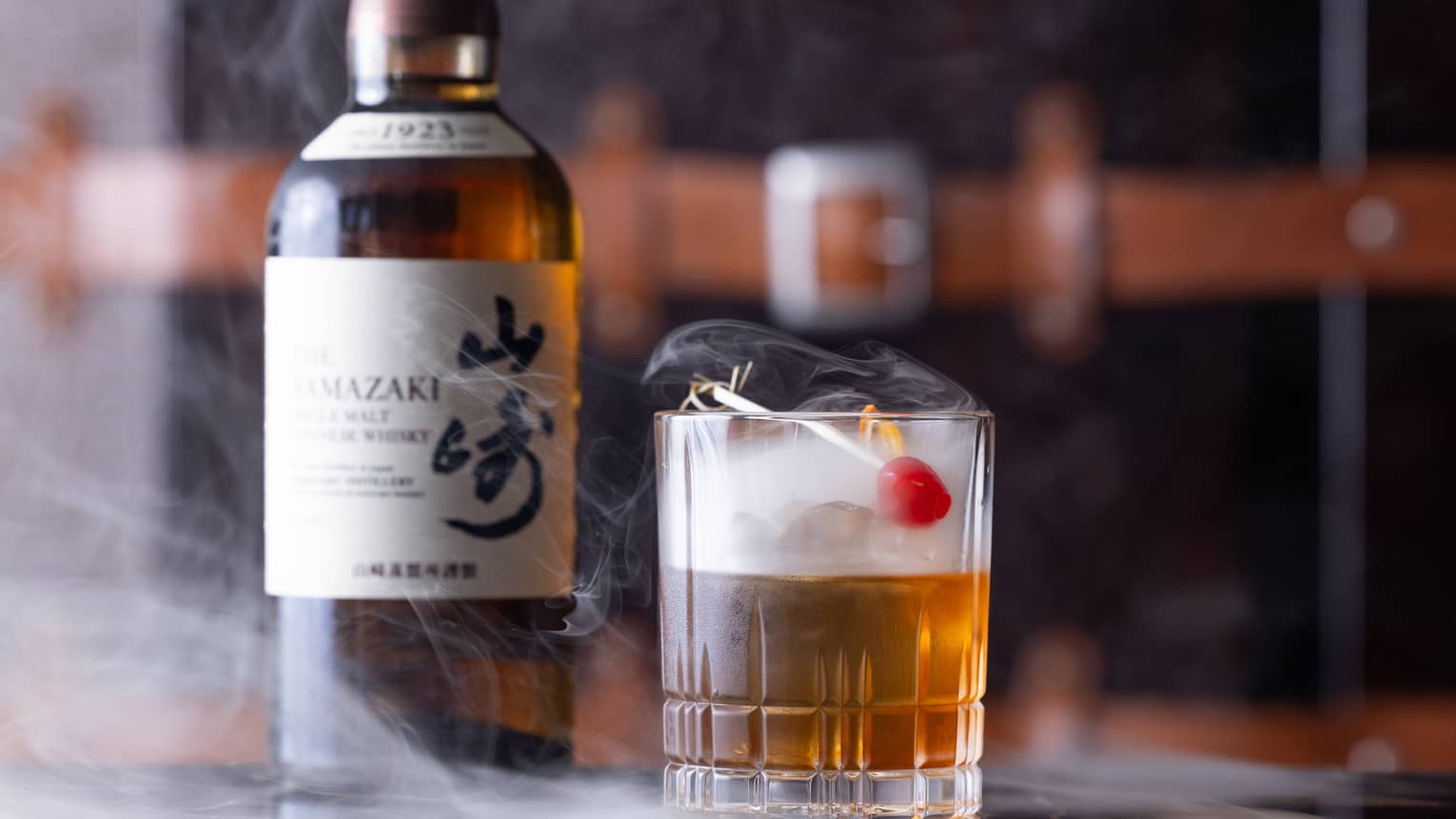 Bottle of Japanese whisky beside half-full cut-crystal glass with red cherry garnish, dry ice mist swirling from the glass.