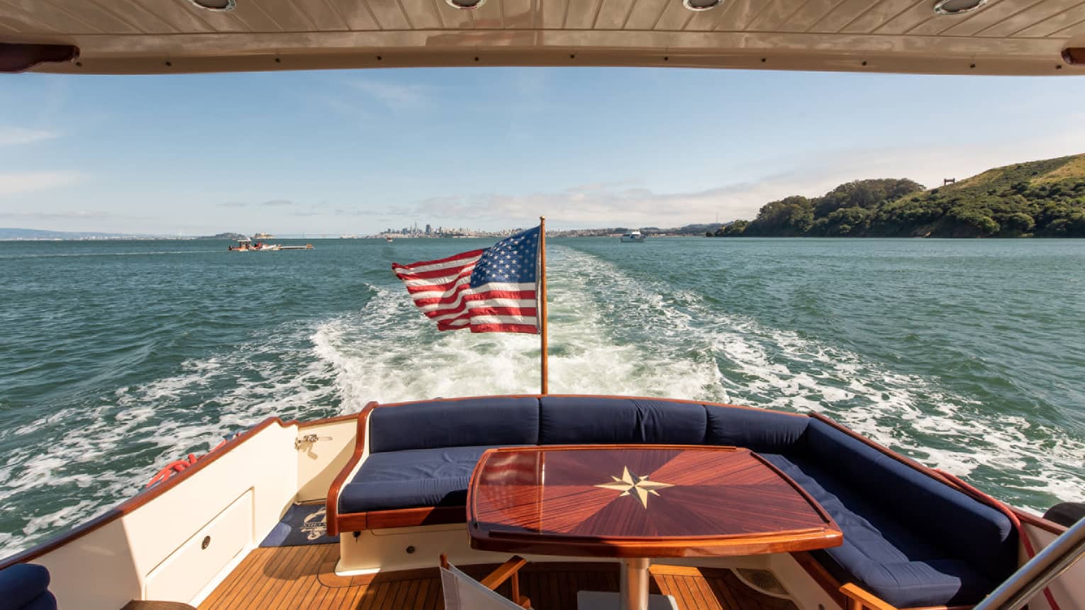 The back of a yacht, with a red table and an American flag, moving along the water.