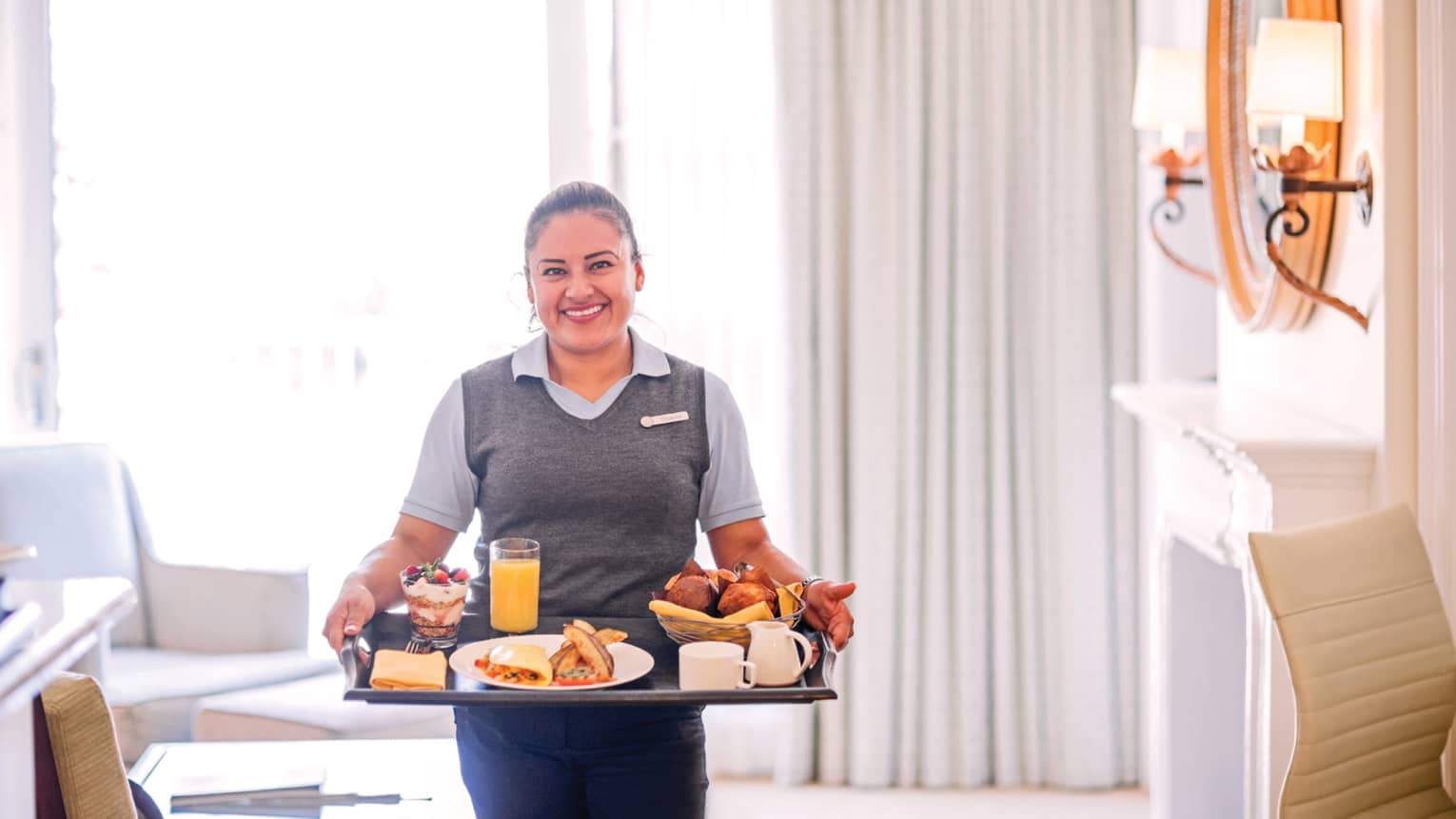 Smiling server carries in-room breakfast tray in elegant white hotel room