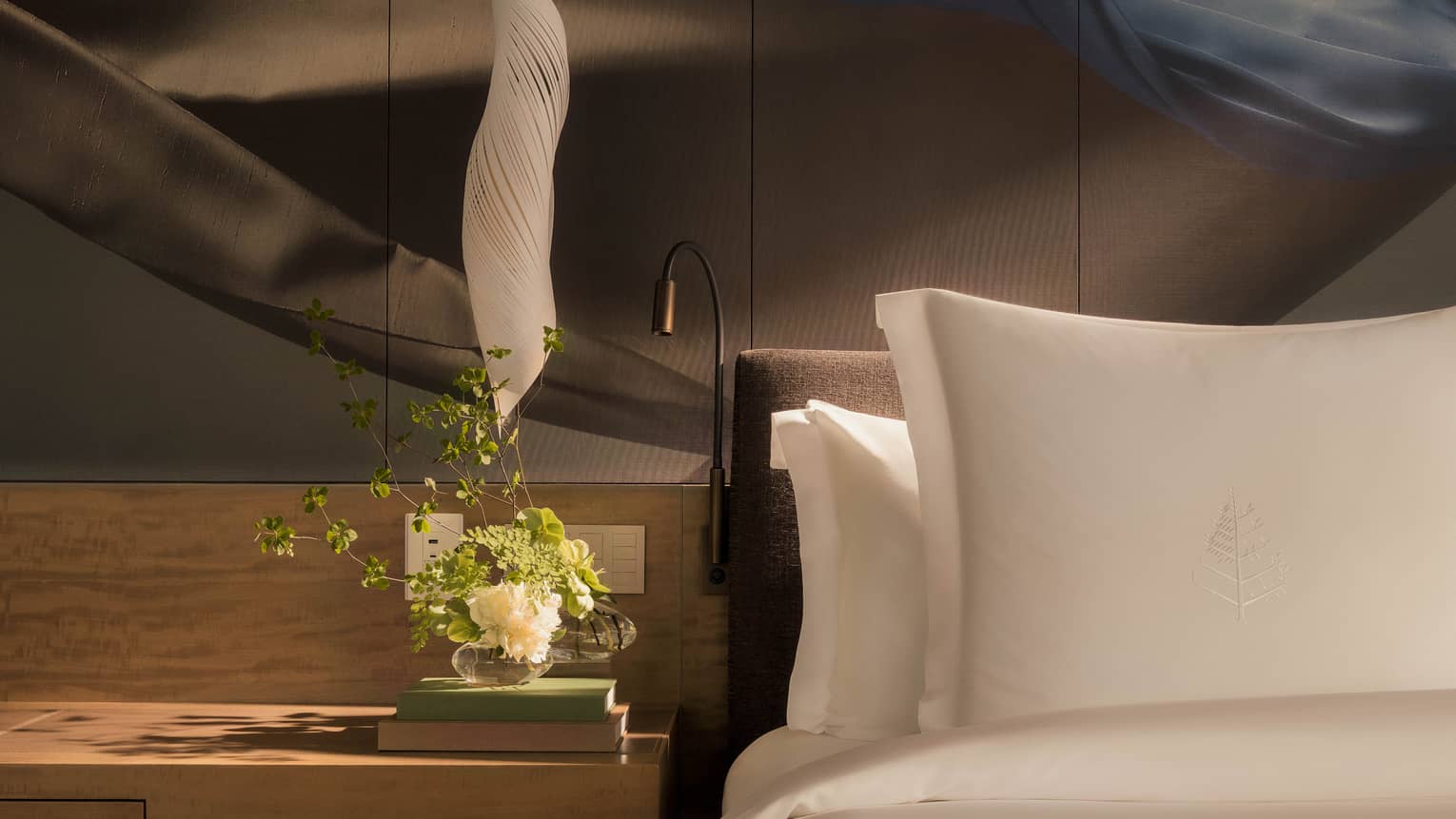 Luxury hotel room bed with artistic accent wall behind white pillows and sheets, and a wooden side table with flowers