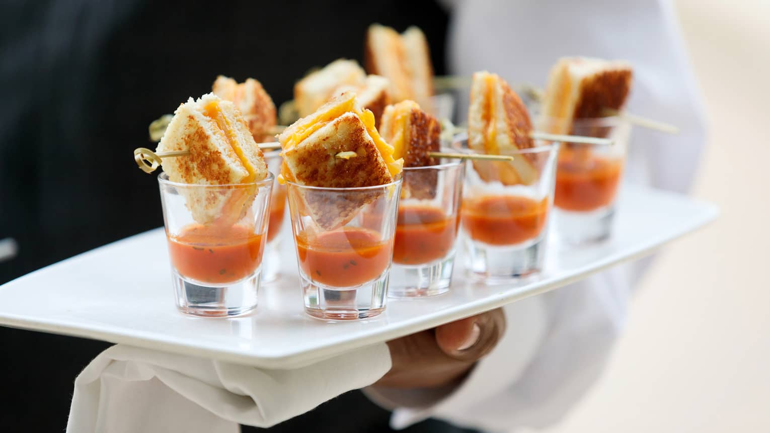 Close up of hotel staff holding platter with tomato soup and small grilled cheese sandwiches in shot glasses
