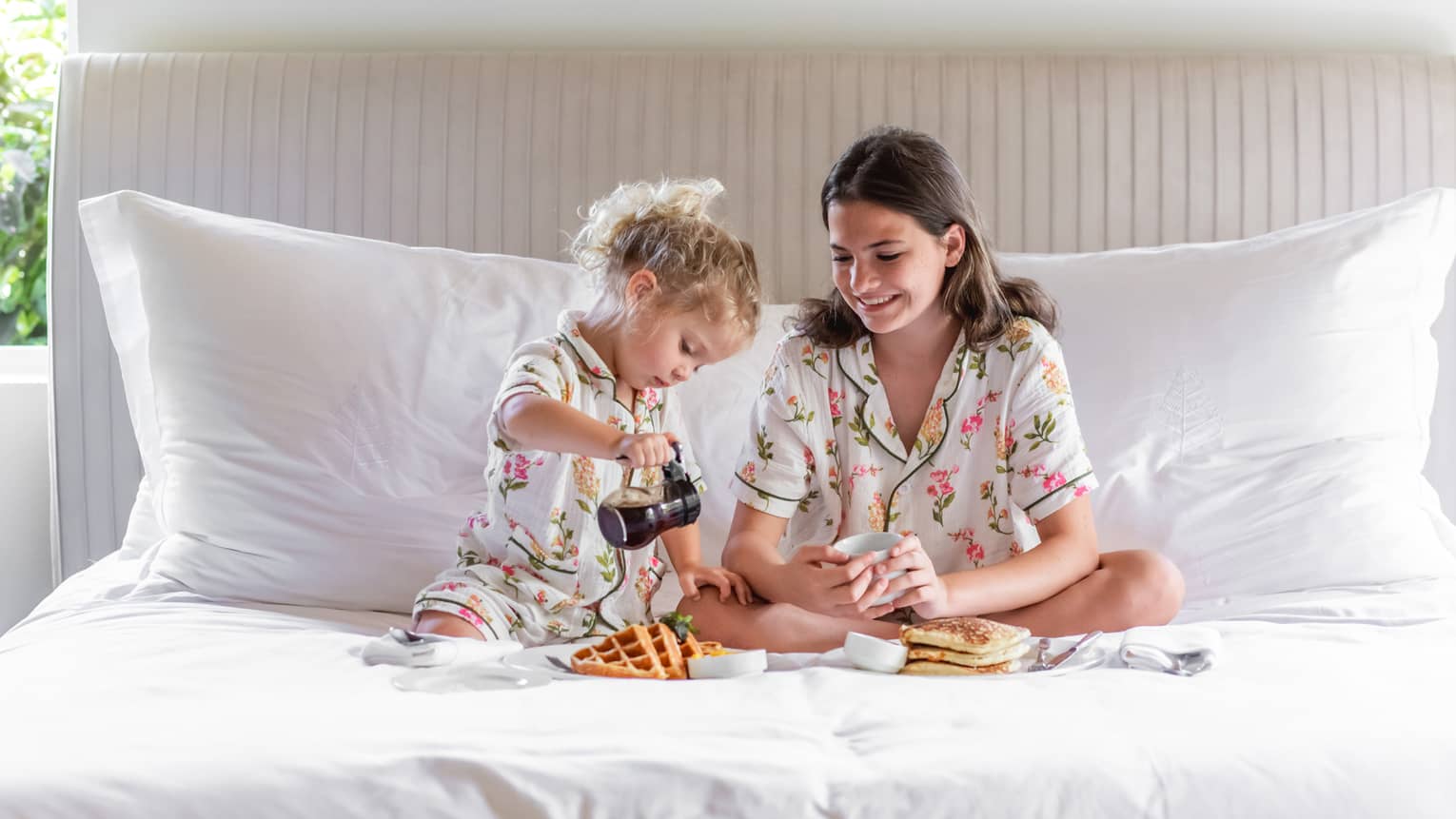 Two young children in matching floral pajamas sit on a white-linen bed eating waffles, as one child purs syrup on their plate