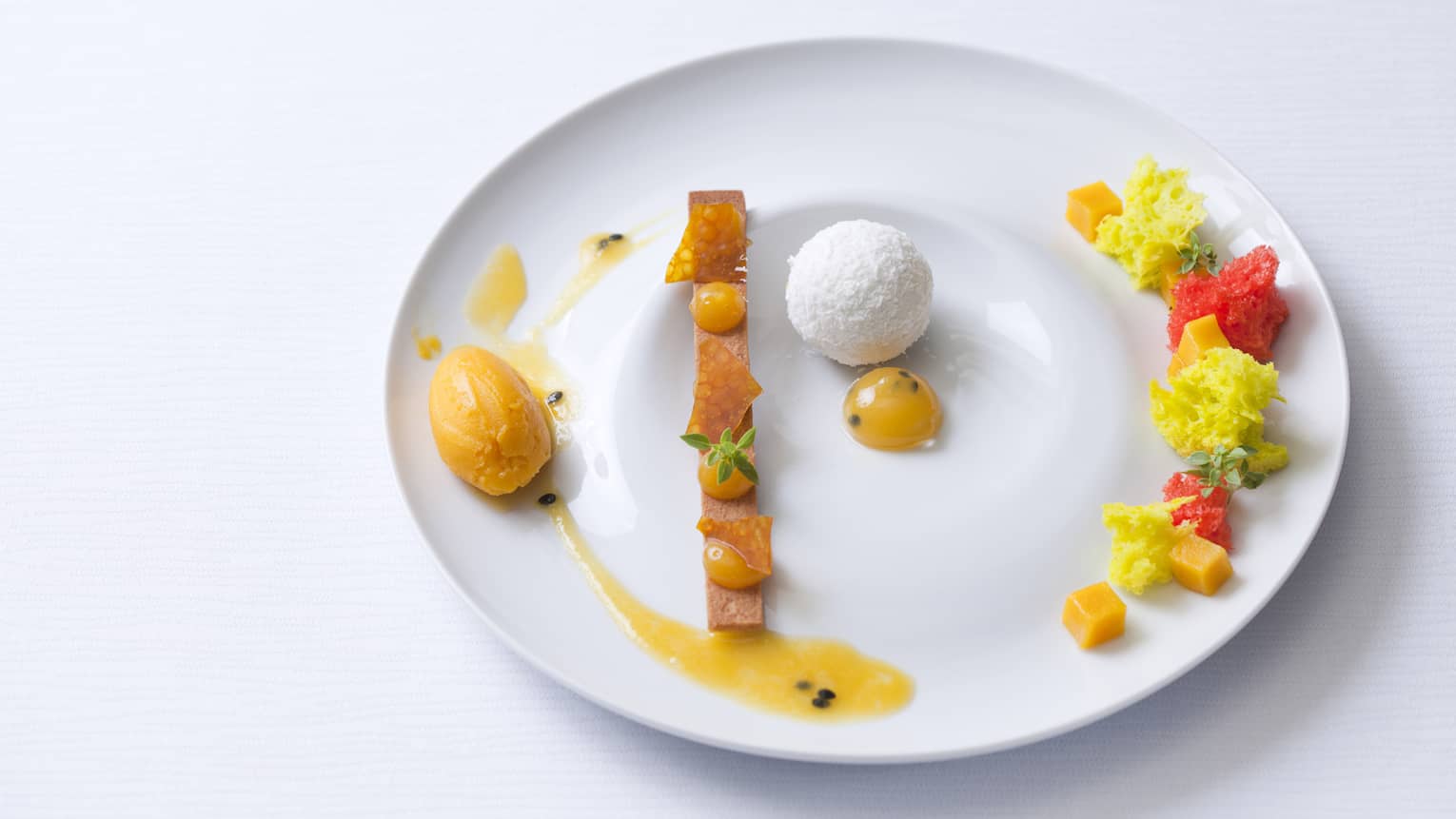 Vanilla sable, passion fruit sauce, coconut mousse ball with passion cremeux insert, marinated fresh mango in sage syrup and mango sorbet on a white plate