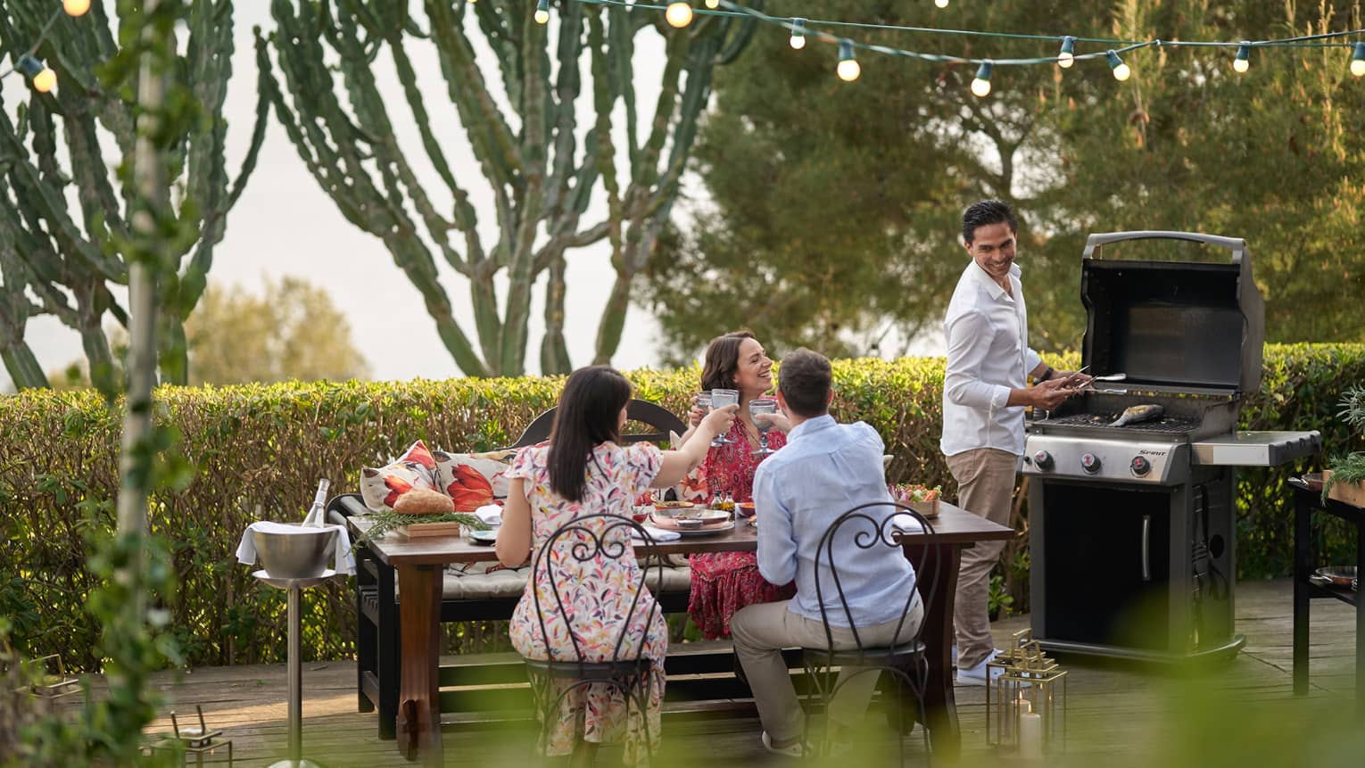 A group of four people grilling food outside at a large table while wearing nice beach clothes.