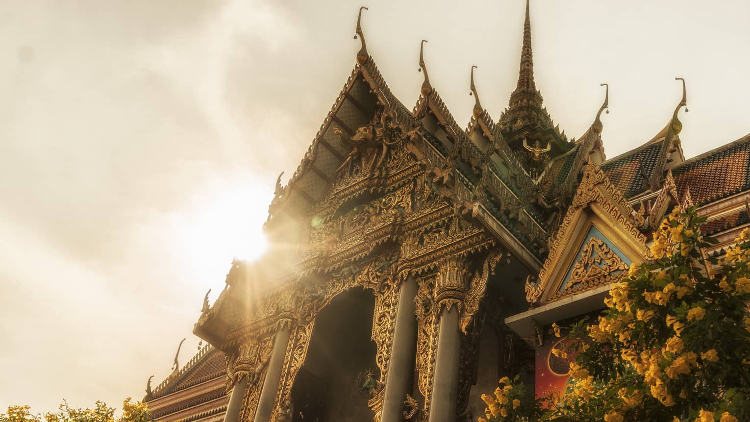 Intricately designed temple with ornate carvings and tiered roofs, illuminated by sunlight, with a tree and yellow flowers in the foreground
