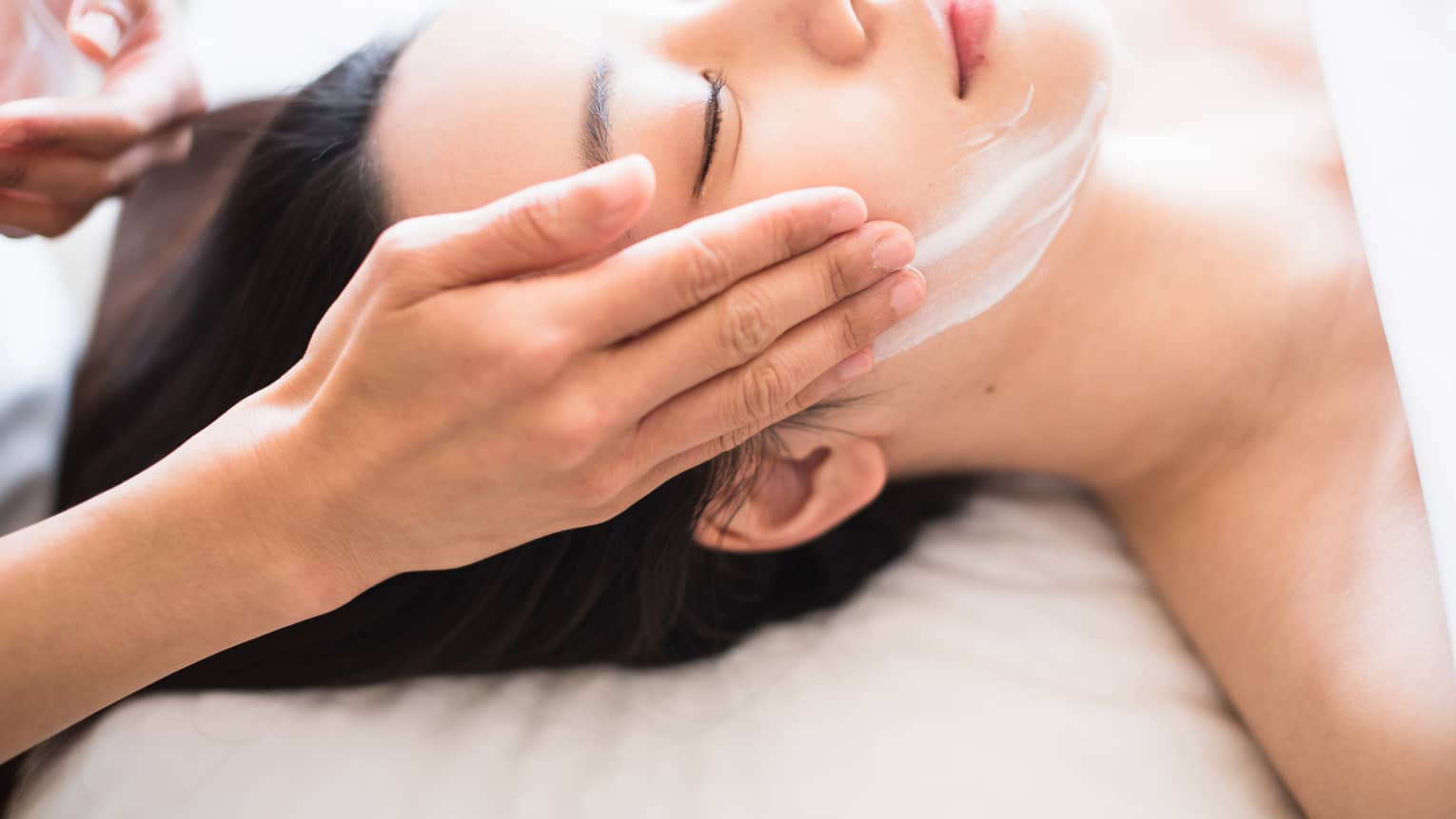 Close-up of spa attendant's hands rubbing white cream on woman's face as she lies on massage table