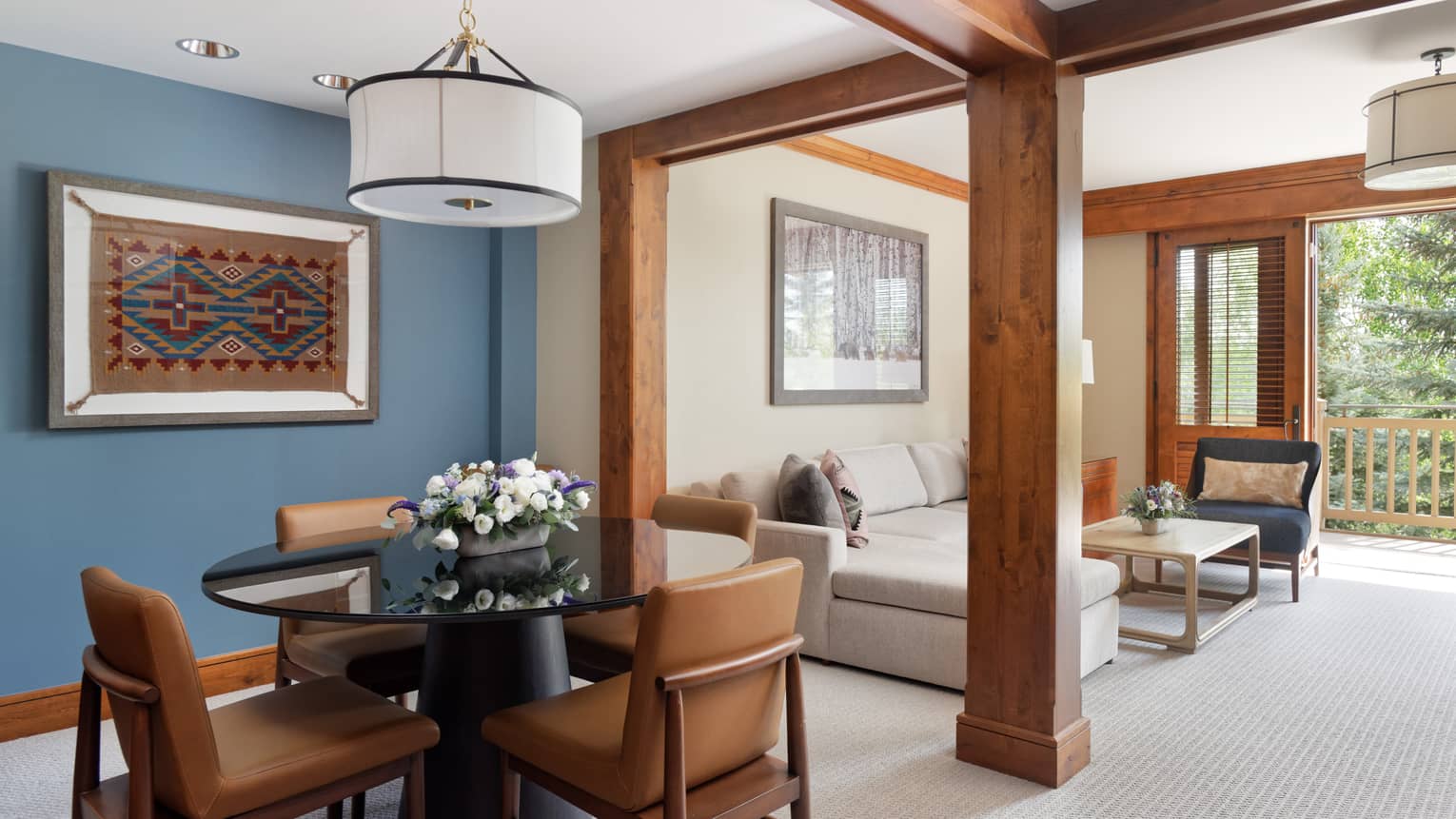 Round dining table with four tan leather chairs, blue accent wall, wooden column separating a light-filled living room