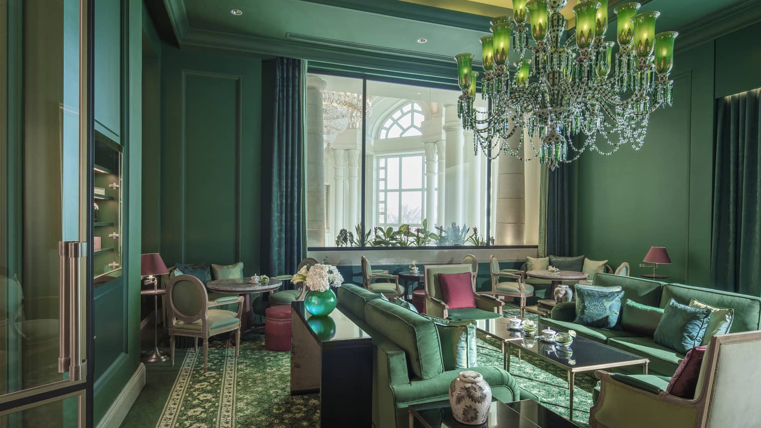 A forest green sitting room with over a dozen velveteen chairs, and a crystal chandelier. 