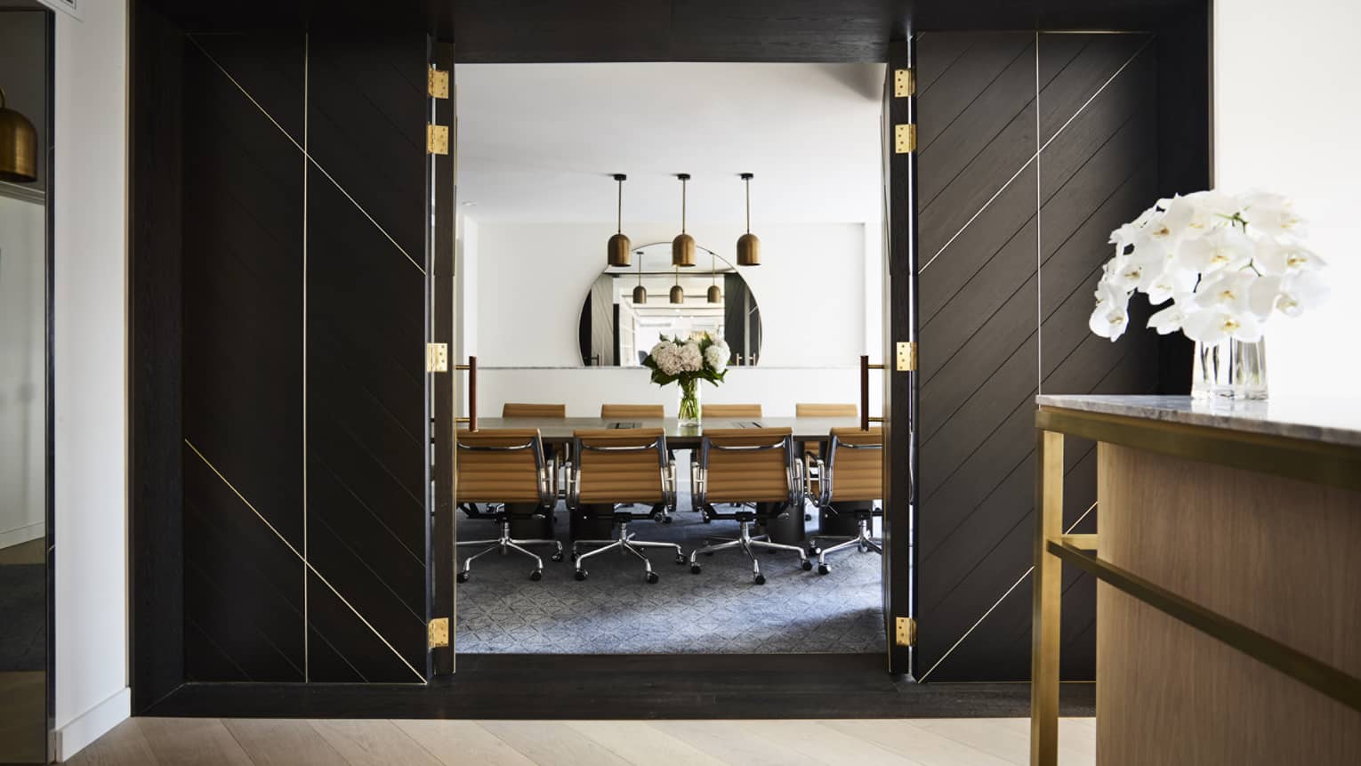 Tall open doors to private boardroom table, swivel chairs under three lights, mirror