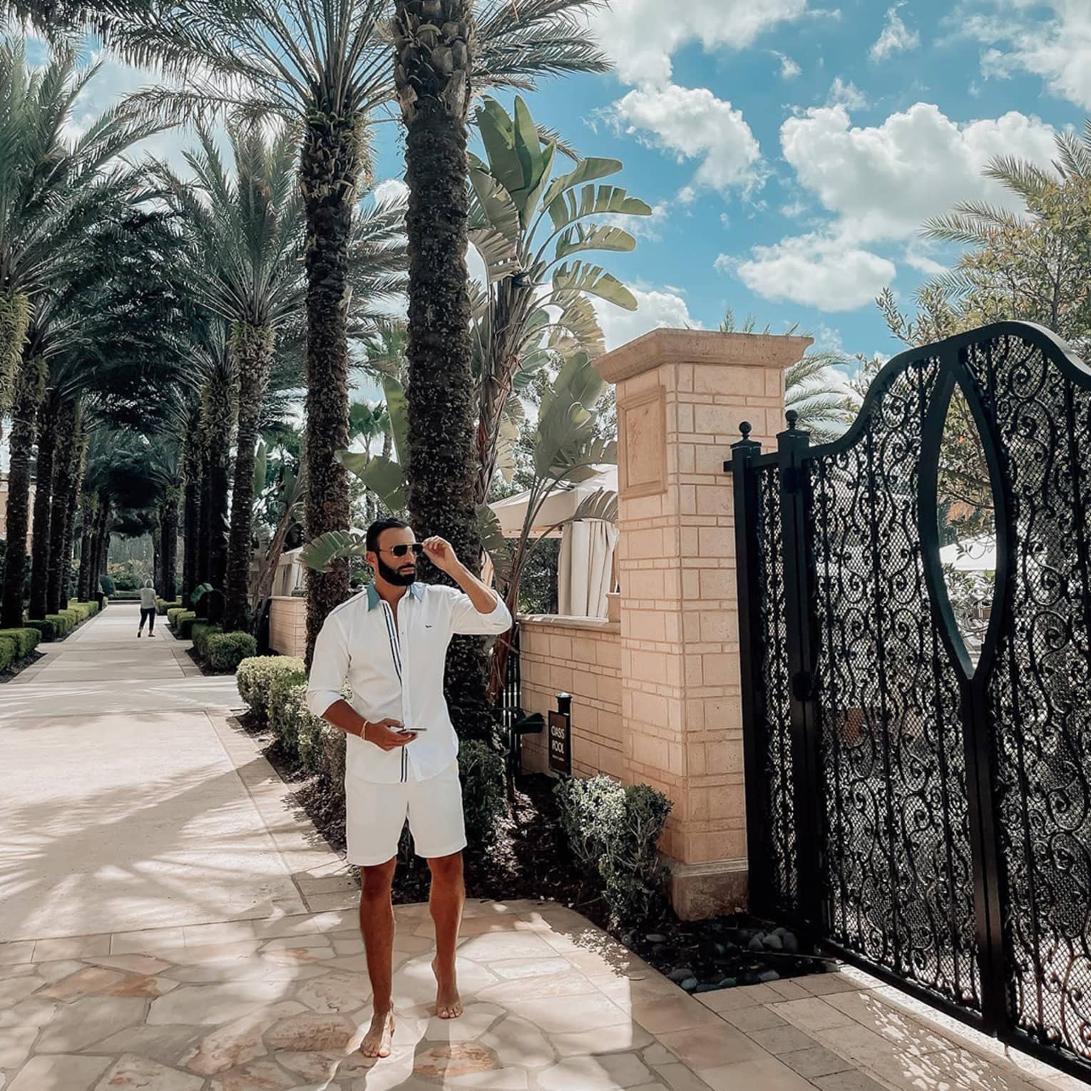 Man wearing white shirt and shorts touches sunglasses by iron gate on sunny path