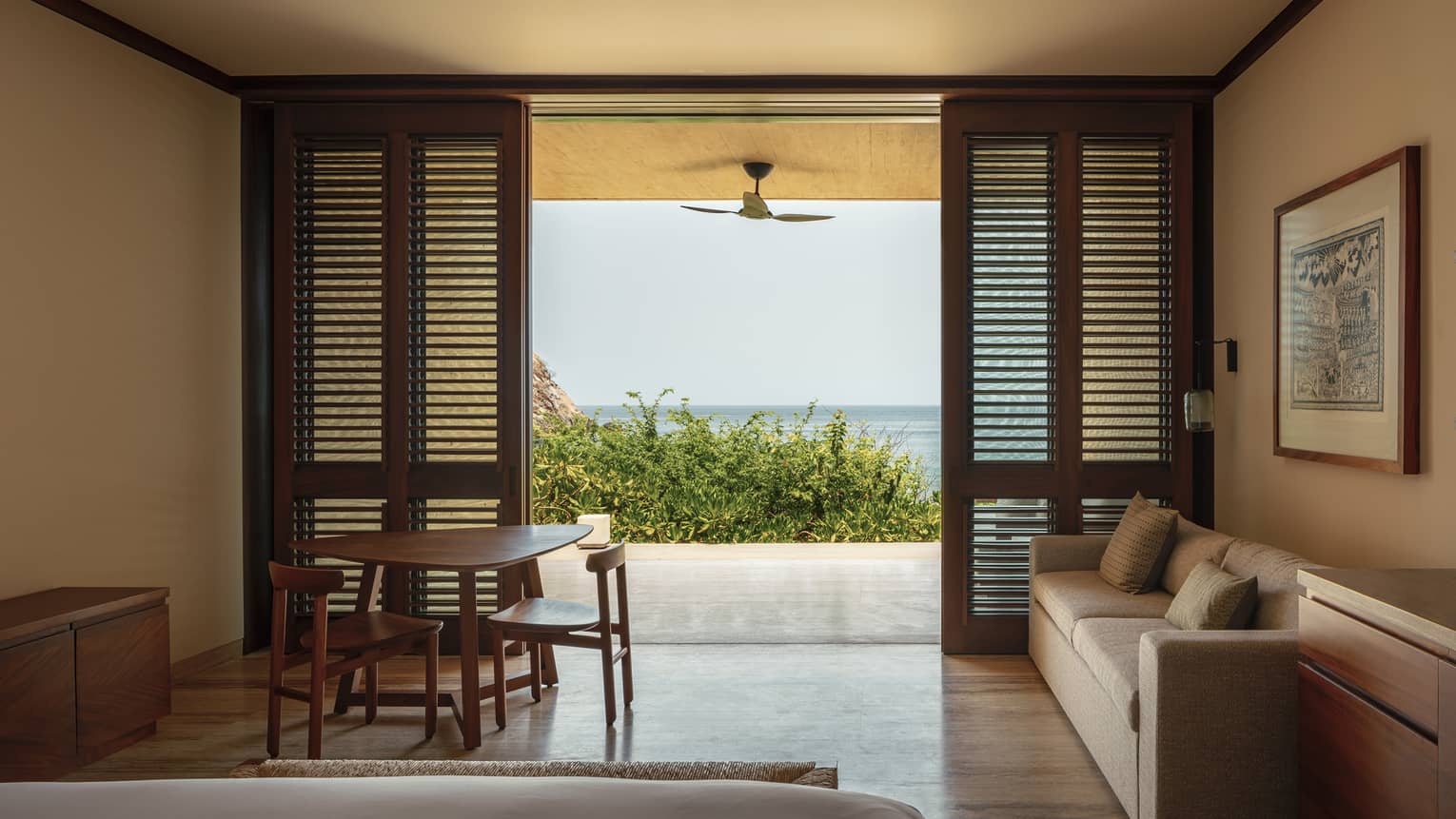 Cozy bedroom with wooden shutters, a sofa and a small dining table. The room opens to a private terrace with ocean views, framed by greenery and a ceiling fan.