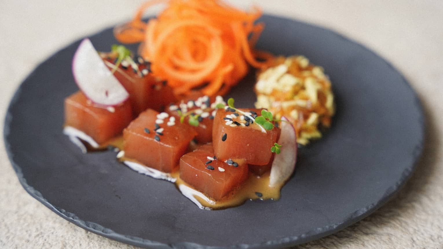 A black plate holds cubes of reddish fish topped by and next to colorful garnishes.