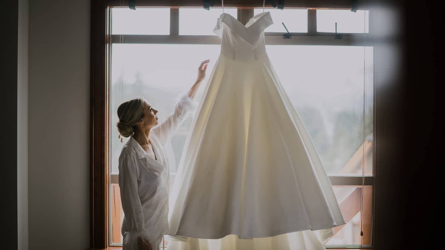Person reaches hand up to touch wedding dress hanging in front of window