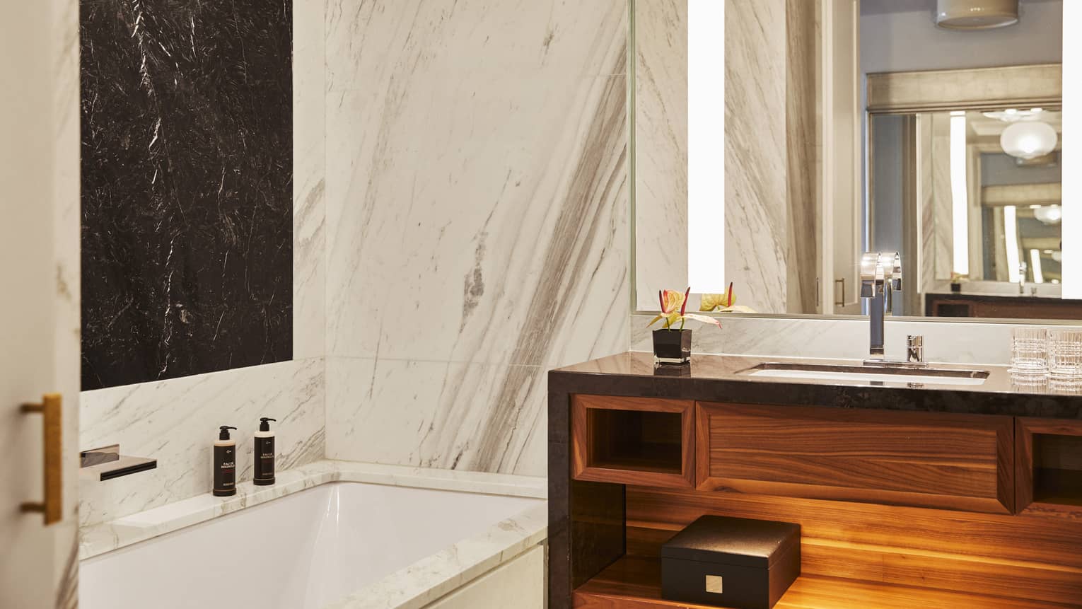 Luxury hotel bathroom with marble walls, black accent panel, built-in bathtub, wooden vanity and large mirror with lighting