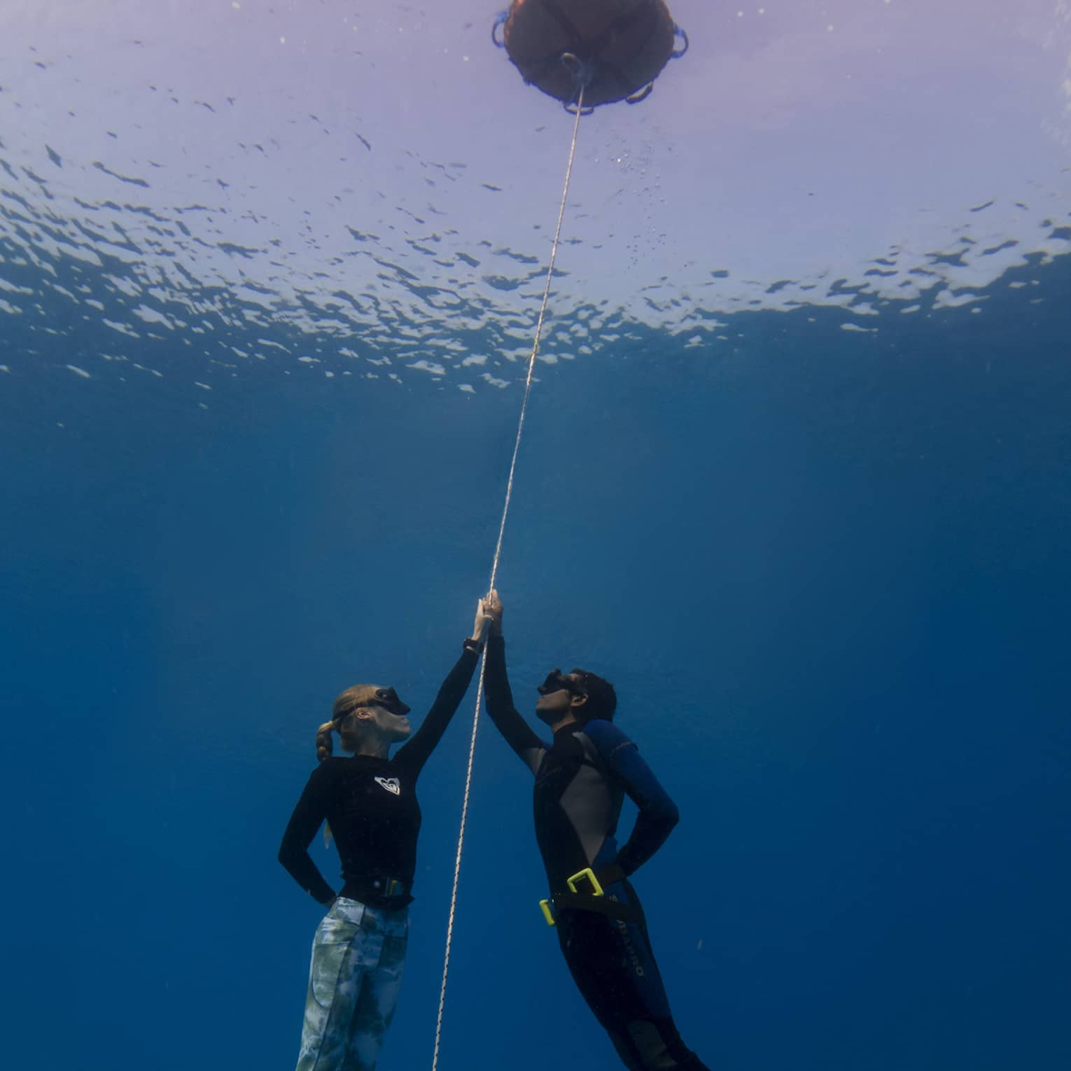 Underwater view of two free-divers, wearing masks and holding onto a rope that attaches to a buoy at the water's surface.