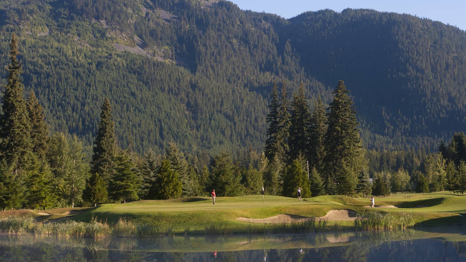 Distant golfers on a green, a pond in front, evergreens behind, dwarfed by a treed mountain filling most of the frame beyond.