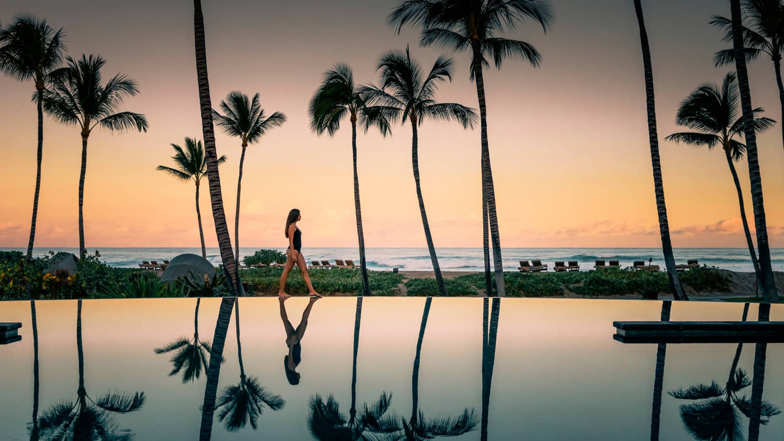 Woman in swimsuit walks by Palm Grove Pool at sunset, palm trees reflected on surface