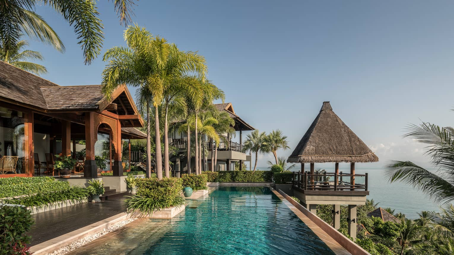 Long outdoor swimming pool under tall palm trees, thatched roof gazebo