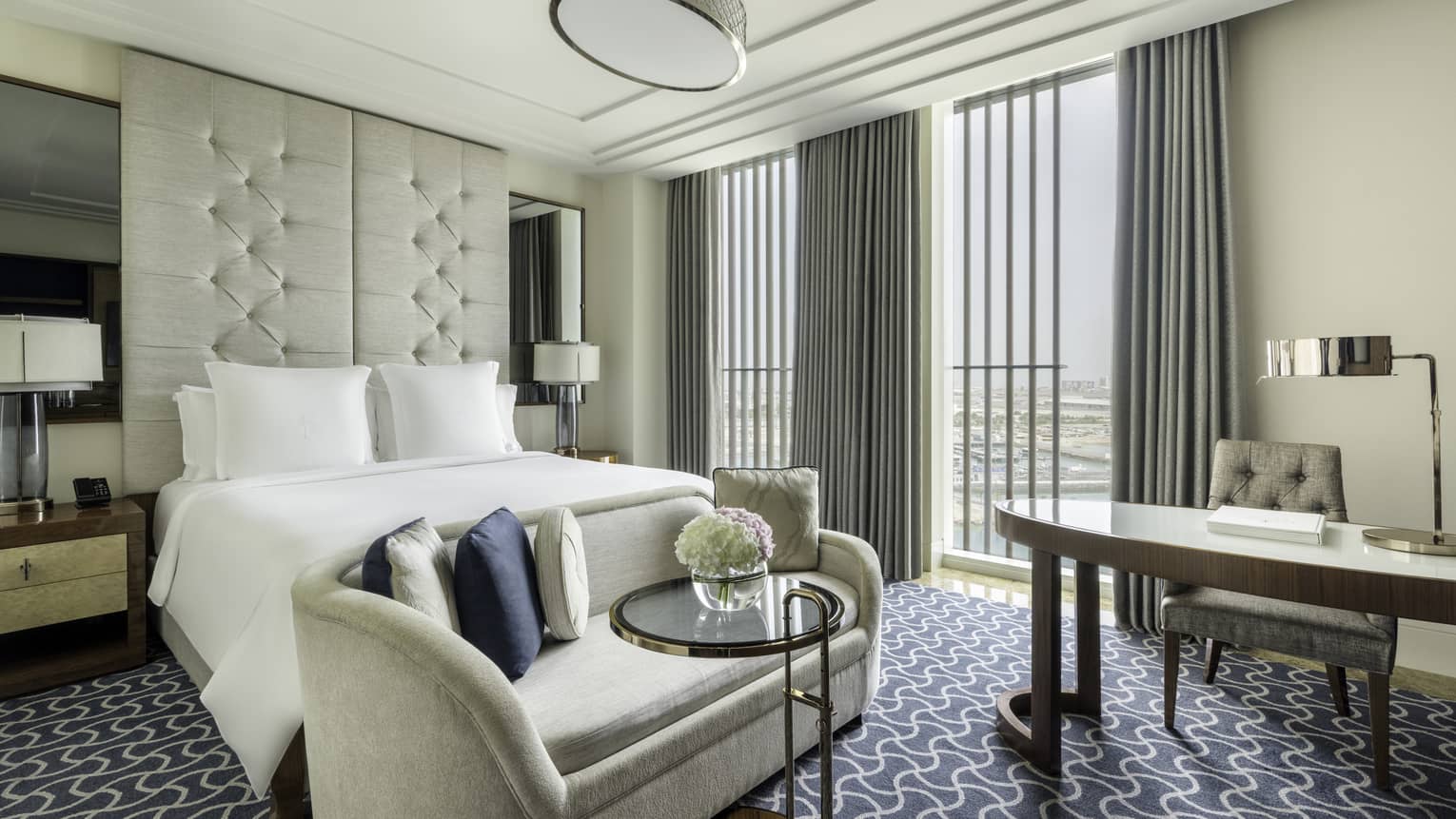 Hotel room with king bed, ceiling-height upholstered headboard, beige chaise, desk and floor-to-ceiling windows