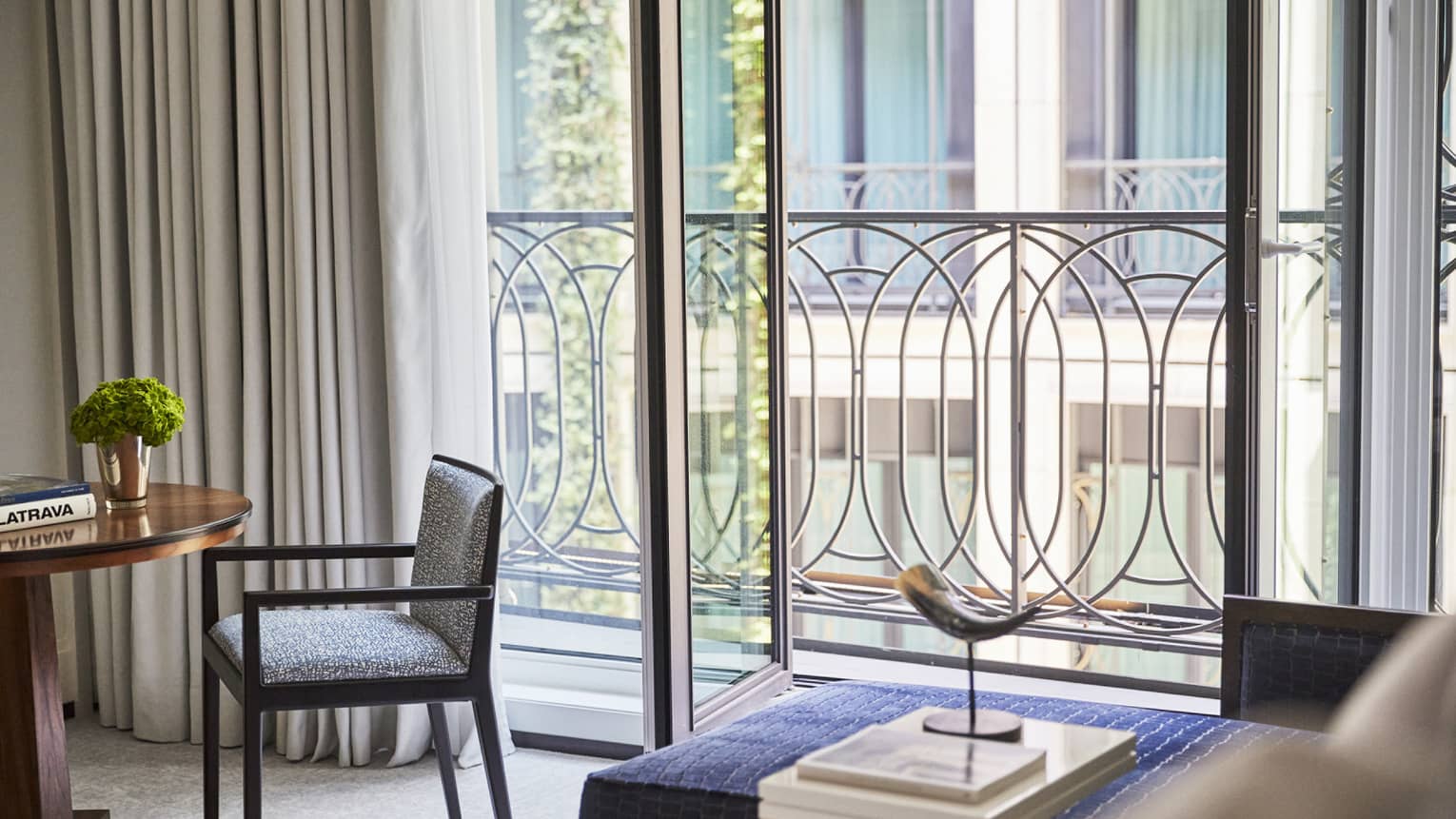 Luxury hotel room interior with a small round table, green plant, chair, horn sculpture, books and decorative railing outside a large window with curtains