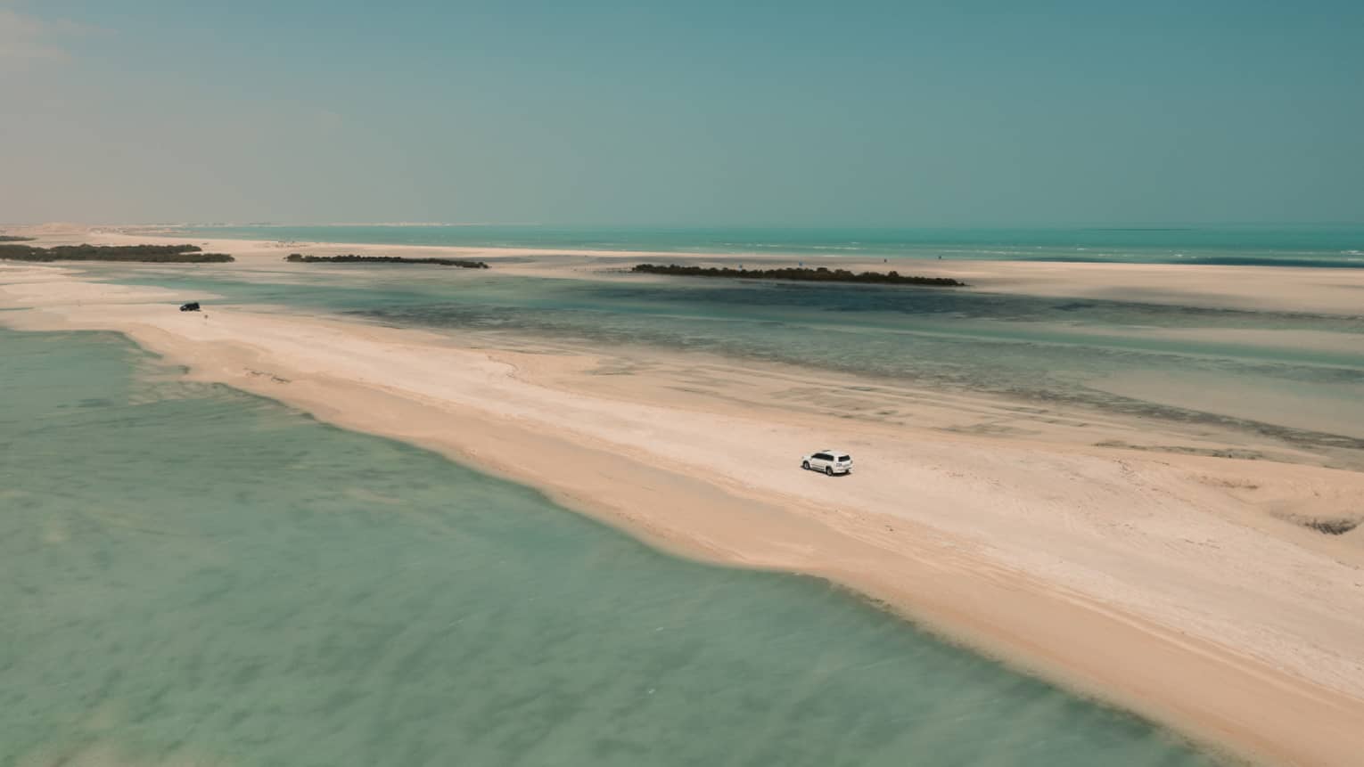 Aerial view of a 4x4 car traversing a sandbar surrounded by shallow turquoise water on either side and a cloudless sky above.