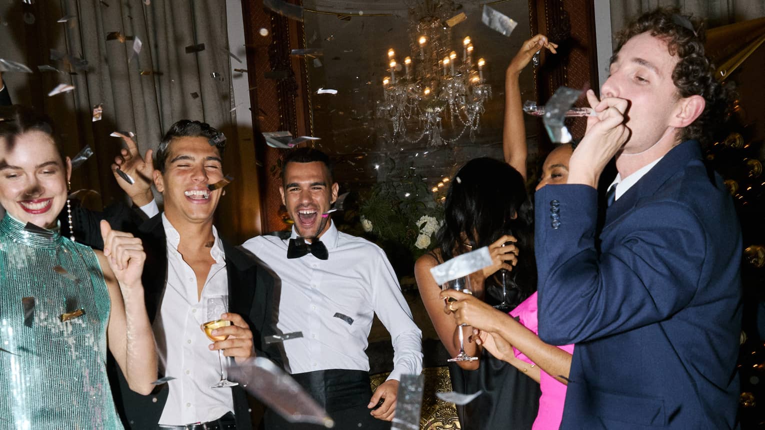 Six people dressed in cocktail party attire smile and celebrate as metallic confetti falls over them in a hotel suite